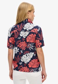 Short-sleeve, button-up shirt with a navy background featuring large red and white tropical leaf patterns and floral accents.