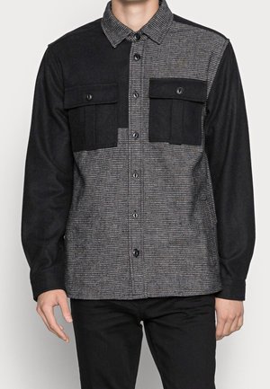 Chemise homme à manches longues avec un bloc de couleurs texturé noir et gris, boutonnière devant, deux poches poitrine noires avec rabats à boutons.