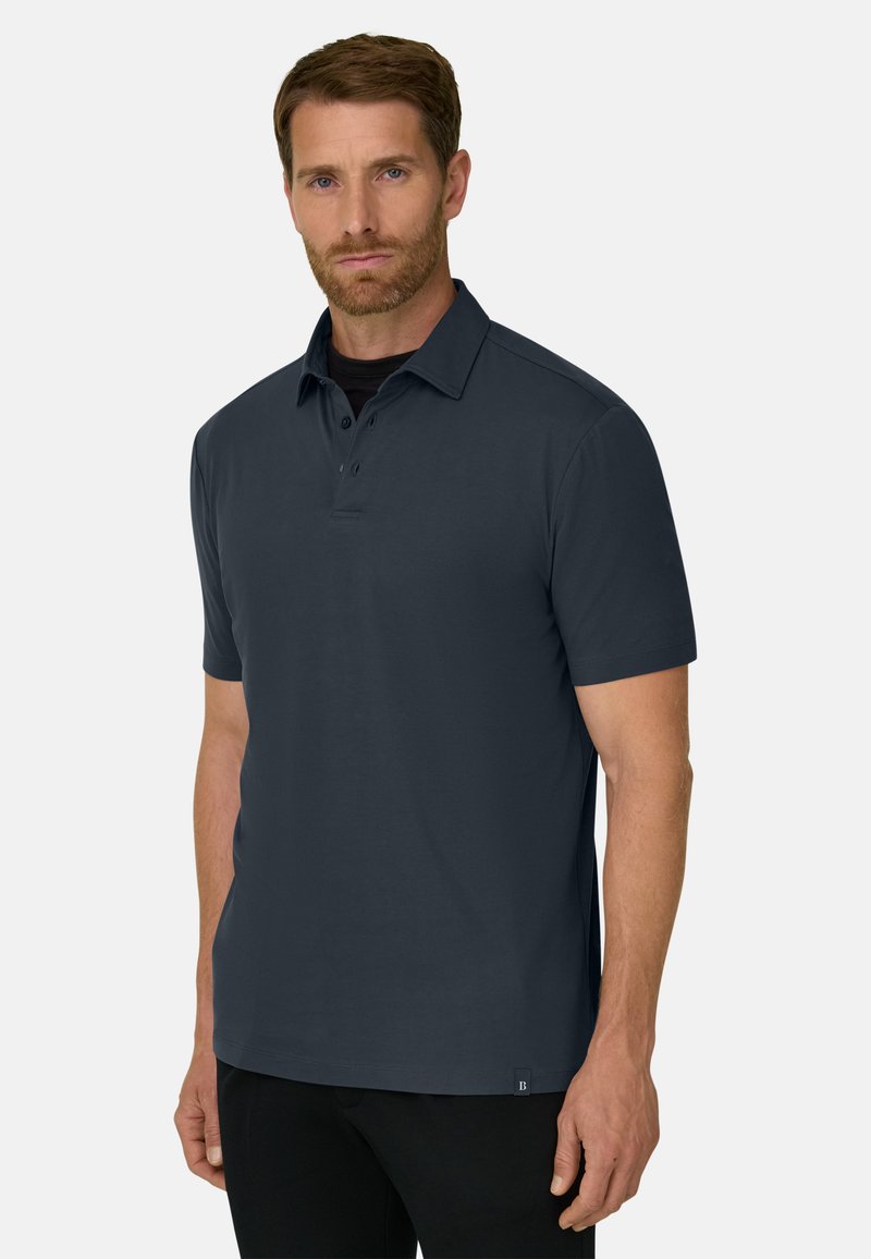 Homme aux cheveux bruns courts et à la barbe, portant un polo bleu foncé à manches courtes et un pantalon noir, debout devant un fond blanc uni.