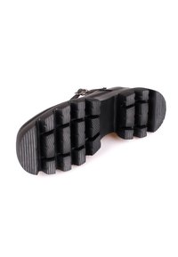 Semelle d'une chaussure noire avec des crampons profonds et larges, et une sangle à boucle sur la partie supérieure, vue en contre-plongée.