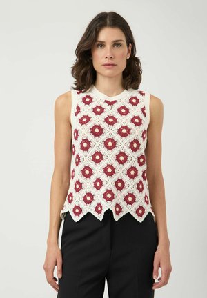 Kobieta o brązowych włosach do ramion nosi białą, bezrękawnikową bluzkę na szydełku z czerwonym wzorem kwiatowym i falowanym wykończeniem, zestawioną z czarnymi spodniami.