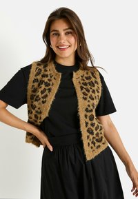 Chaleco marrón de felpa con estampado de leopardo negro, con un diseño abierto. Llevado sobre una camiseta negra, combinado con una falda negra.