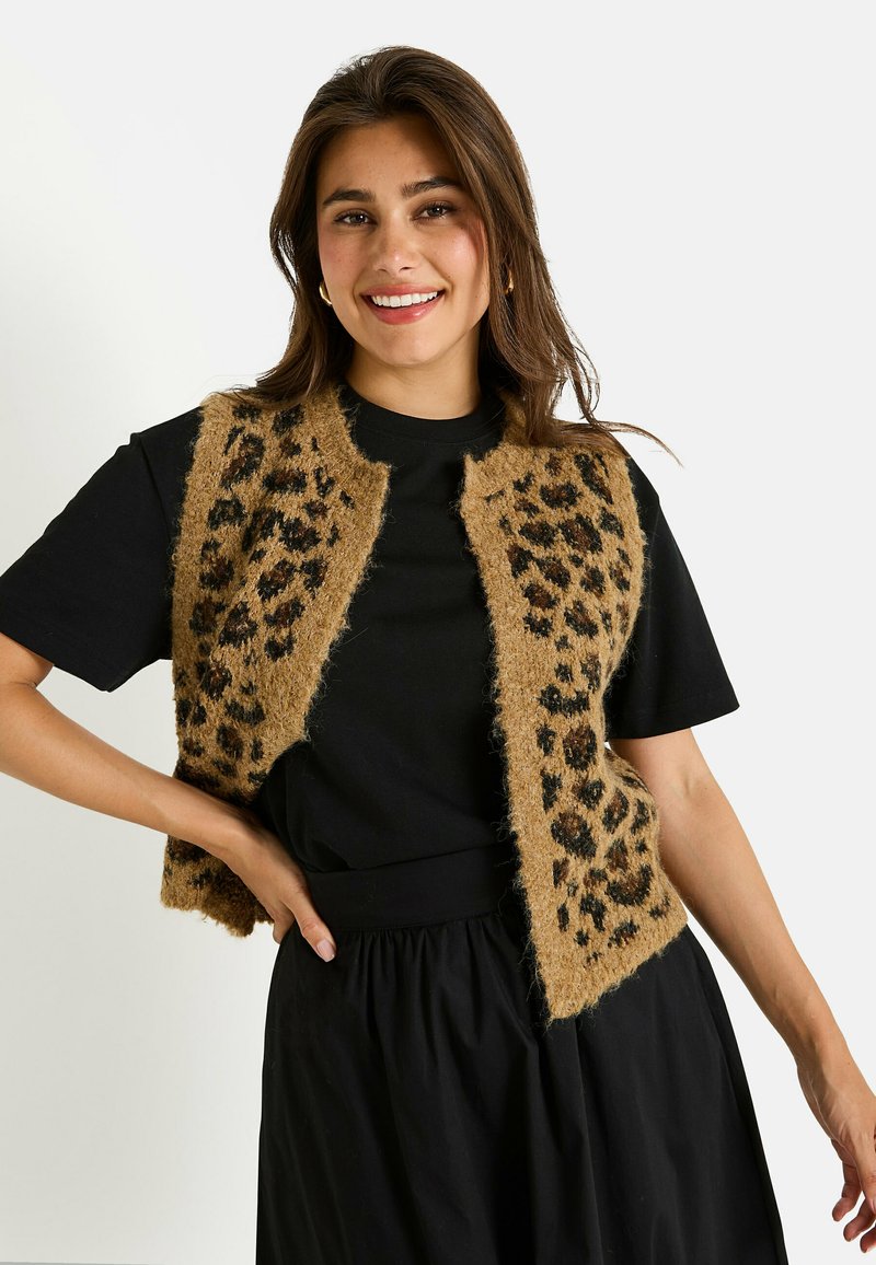 Chaleco marrón de felpa con estampado de leopardo negro, con un diseño abierto. Llevado sobre una camiseta negra, combinado con una falda negra.