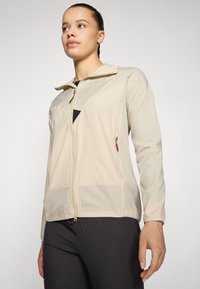 Veste à zip beige clair avec capuche, dotée d'un tissu texturé, de poches latérales et de détails en maille le long des manches.