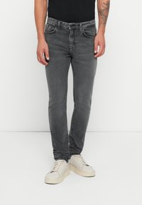 Szürke, slim-fit farmernadrág, enyhén kopott mosással, öt zsebes dizájnnal és öv hurkokkal, tört fehér sportcipővel egy semleges háttér előtt.