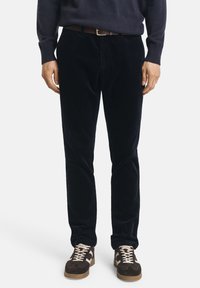 Pantalon en velours côtelé bleu marine à coupe slim, avec une texture fine. Porté avec un pull sombre et des baskets brunes avec des accents clairs.