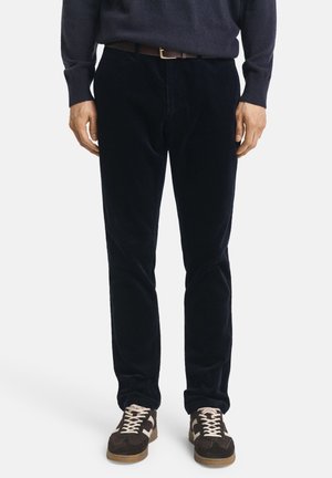 Pantaloni in corduroy blu navy con una vestibilità slim, caratterizzati da una fine trama. Indossati con un maglione scuro e sneaker marroni con dettagli chiari.