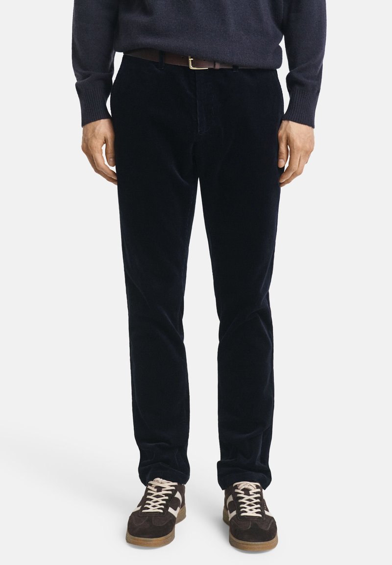Pantalon en velours côtelé bleu marine à coupe slim, avec une texture fine. Porté avec un pull sombre et des baskets brunes avec des accents clairs.