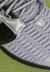 Close-up de um sapato atlético cinza com tecido de malha tricotada, sola preta e uma fita metálica prateada sobre os atacadores, colocado sobre uma superfície texturizada verde.