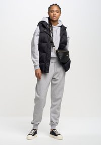 Schwarze Steppweste über einem grauen Hoodie und passenden Sweatpants. Eine schwarze Umhängetasche und schwarz-weiße Sneakers vervollständigen das Outfit.