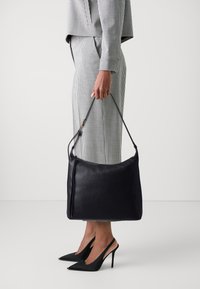 Schwarze Lederhandtasche mit glatter Textur, ausgestattet mit einem langen Riemen und subtilen Reißverschlussdetails. Das Modell trägt einen karierten Anzug und schwarze spitze Absätze.