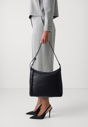 Schwarze Lederhandtasche mit glatter Textur, ausgestattet mit einem langen Riemen und subtilen Reißverschlussdetails. Das Modell trägt einen karierten Anzug und schwarze spitze Absätze.