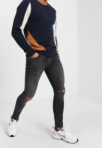 Felpa navy con pannelli crema e marrone, abbinata a jeans neri svaligiati e scarpe da ginnastica bianche con accenti neri.