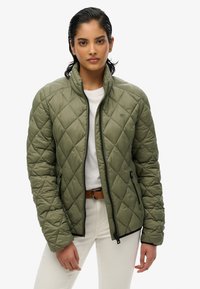 Groene gewatteerde jas met opstaande kraag, ritssluiting aan de voorkant en twee zijzakken. Draagt over een wit shirt en gecombineerd met lichte broeken.