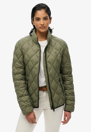 Veste matelassée verte avec un col montant, fermeture éclair frontale et deux poches latérales. Portée sur une chemise blanche et associée à un pantalon clair.