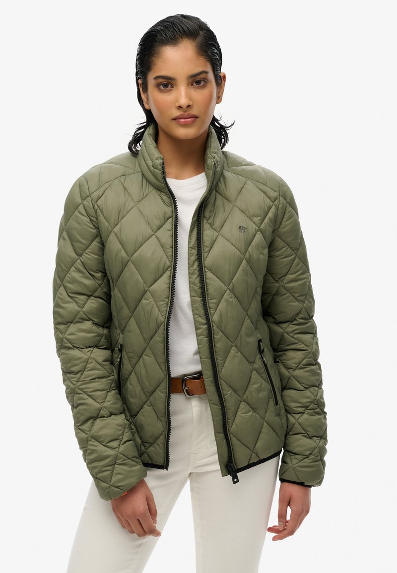 Groene gewatteerde jas met opstaande kraag, ritssluiting aan de voorkant en twee zijzakken. Draagt over een wit shirt en gecombineerd met lichte broeken.