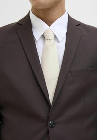 Abito marrone con un singolo bottone nero, camicia bianca e cravatta crema chiara; texture liscia e vestibilità sartoriale in evidenza.