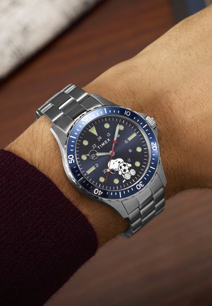 Ρολόι χειρός με ασημένιο μεταλλικό λουράκι και μπλε καντράν, που δείχνει τη μάρκα Timex και τον Snoopy να κρατά μια ποδοσφαιρική μπάλα στην επιφάνεια του ρολογιού.