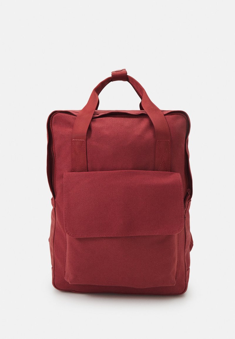 Sac à dos en toile rouge avec deux bretelles, une poche avant à rabat et une forme rectangulaire. Texture lisse et détails de quincaillerie minimalistes.