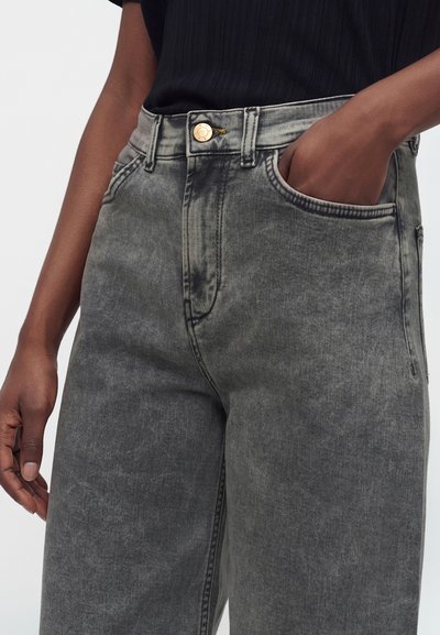 Personne portant un jean noir délavé taille haute, une main dans la poche avant et un haut côtelé noir.