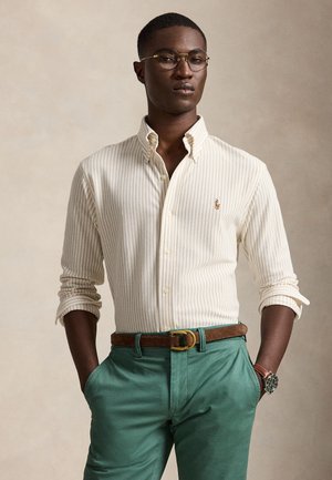Homme avec des lunettes portant une chemise beige à rayures boutonnée, un pantalon vert, une ceinture marron et une montre-bracelet, debout les mains dans les poches.