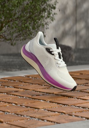 Bílá sportovní bota s fialovou podrážkou a černými detaily levitující nad dřevěným lamelovým povrchem venku poblíž betonové zdi a zeleně.