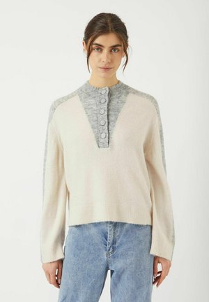 Femme portant un pull en tricot crème et gris clair avec des boutons sur la poitrine, associé à un jeans bleu clair, se tenant contre un fond blanc.