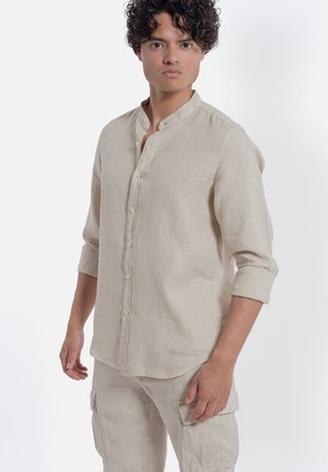 Giovane uomo con capelli ricci che indossa una camicia beige a maniche lunghe con bottoni e pantaloni cargo abbinati, in piedi davanti a uno sfondo bianco.