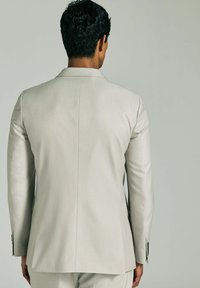 Giacca elegante beige chiaro con una texture liscia, apertura posteriore singola e dettagli in bottoni neri. Fit su misura con spalle strutturate e maniche lunghe.