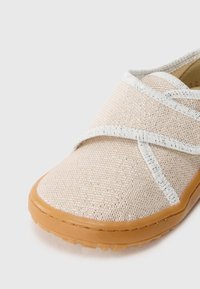 Zapatilla slip-on de color beige claro con un upper texturizado y tejido, y acentos plateados. Presenta una suela de goma flexible y un diseño de tiras cruzadas.