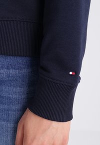 Felpa blu navy con polsini a coste, dotata di un piccolo logo patch tricolore sulla manica. Texture in denim visibile sullo sfondo.