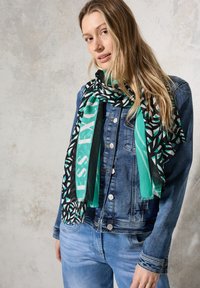 Rektangulär scarf i teal, svart och vitt med geometriska mönster och text. Lättviktsmaterial med fransade kanter, bärs över en denimjacka.