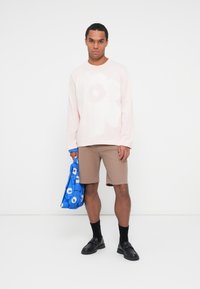 Camicia a maniche lunghe rosa chiaro con una grande stampa di fiori bianchi, abbinata a pantaloni corti beige e a una borsa a tracolla floreale blu. Scarpe e calze nere.