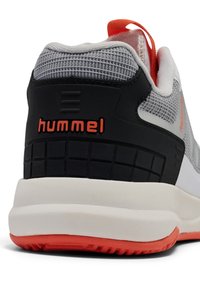 Tacón de zapatilla deportiva gris y blanco con suela negra y naranja, parte superior de malla y logo "hummel" en color naranja en el contrafuerte negro.