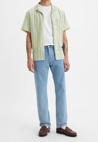 Kortärmad skjorta med knappar i gula och blå ränder, kombinerad med ljusblå raka jeans och bruna loafers, med vita strumpor.