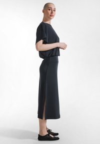 Mujer con cabeza rapada que lleva una blusa negra suelta, falda negra de talle alto con abertura lateral y zapatos planos negros, de pie de perfil.