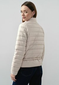 Beżowa kurtka puchowa z poziomymi pikowaniami, wysokim kołnierzem i elastycznym pasem. W połączeniu z ciemnoniebieskimi dżinsami. Gładka tekstura.