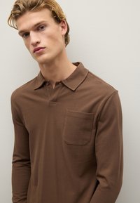 Braunes langärmliges Poloshirt mit Kragen, das über eine Brusttasche und einen strukturierten Stoff verfügt. Einfaches Design mit Knopfdetails am Hals.