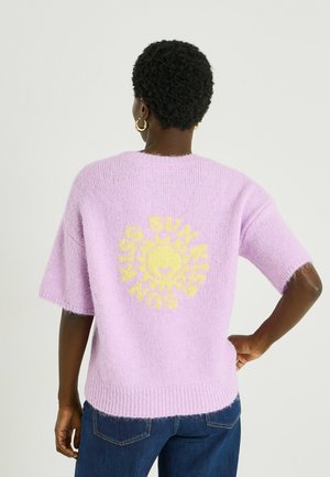 Persona con cabello corto y rizado, lleva pendientes de aro dorados y un suéter de punto lavanda con un sol amarillo y el texto "SUN" en la espalda.