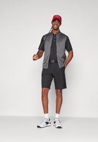 Tenue de golf comprenant un gilet sans manches gris, un polo noir, un short noir et des baskets blanches avec accents noirs. Les accessoires incluent une casquette rouge.
