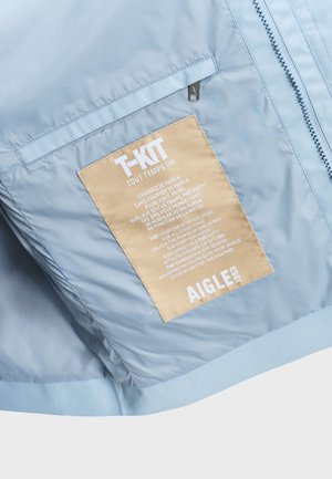 Light blue jacket with beige rectangular patch labeled "T-KIT" and brand name "AIGLE 1853" near a vertical zipper.