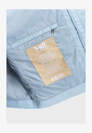 Hellblaue Jacke mit beigem rechteckigem Aufnäher mit der Aufschrift "T-KIT" und dem Markennamen "AIGLE 1853" in der Nähe eines vertikalen Reißverschlusses.