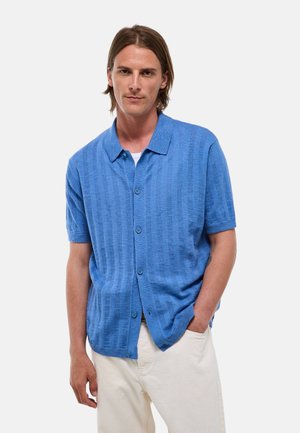 Homme aux cheveux bruns mi-longs portant une chemise bleue à manches courtes boutonnée et un pantalon clair, debout, une main dans la poche.