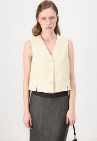Beige kort väst med V-hals, knappstängning fram och sidofickor, i kombination med en mörk kjol med vertikala ränder. Enkla, rena linjer.