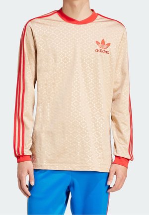 Beżowa koszulka Adidas z długimi rękawami, z czerwonymi paskami na rękawach i logo na piersi, noszona z jasnoniebieskimi spodniami.