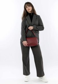 Grau gemischter Wollanzug mit einem taillierten Blazer und weit geschnittenen Hosen. Hält eine kleine rote Ledertasche. Weiße Sneakers runden das Outfit ab.