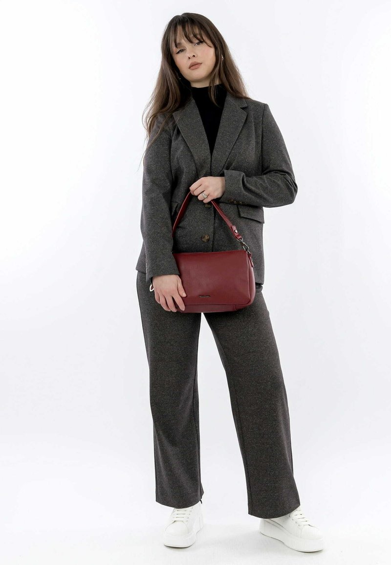 Grau gemischter Wollanzug mit einem taillierten Blazer und weit geschnittenen Hosen. Hält eine kleine rote Ledertasche. Weiße Sneakers runden das Outfit ab.