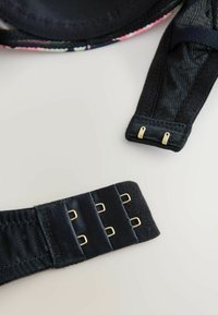 Černý ramínek podprsenky se zlatými dvojitými háčky a černými zapínáními na háček a oko na světlém pozadí.