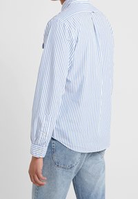 Långärmad skjorta med knäppning i vitt med blå vertikala ränder, med en spetsig krage och enskilda knappar i ärmsluten. Skräddarsydd passform med rundad nederkant.