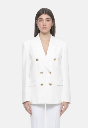 Donna con capelli lisci e scuri che indossa un blazer doppiopetto bianco con bottoni dorati e pantaloni bianchi abbinati, in piedi davanti a uno sfondo neutro.